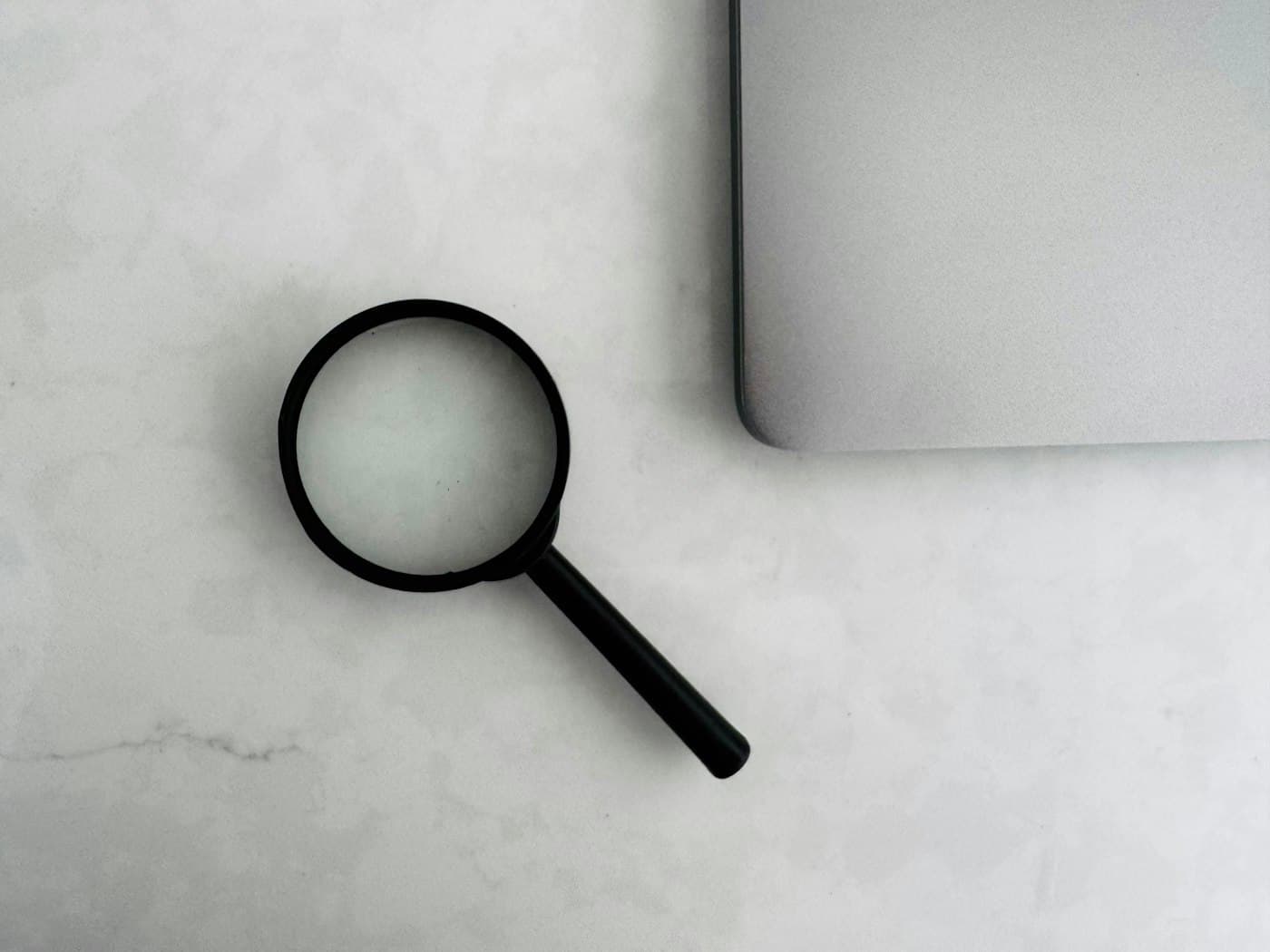 A magnifying glass lying next to the corner of a laptop on a pale stone surface