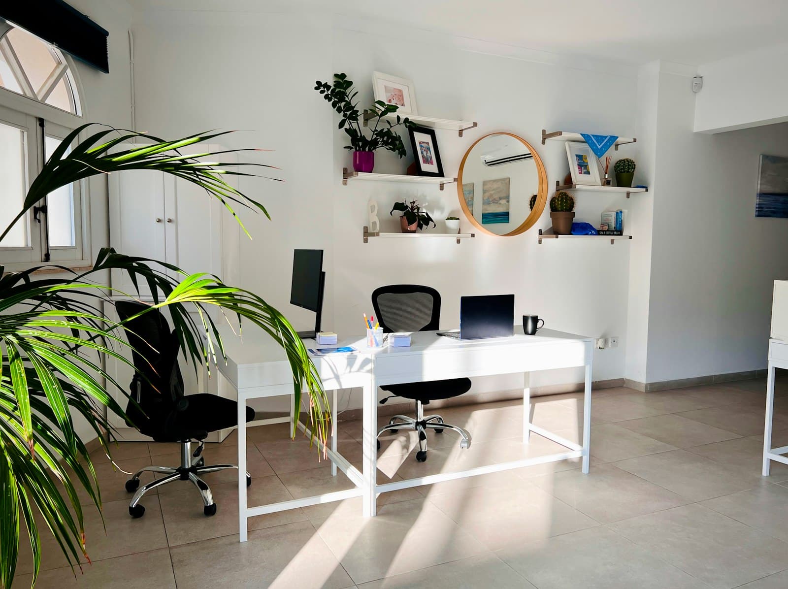 A bright white workspace with two desks, shelves and daylight across the floor