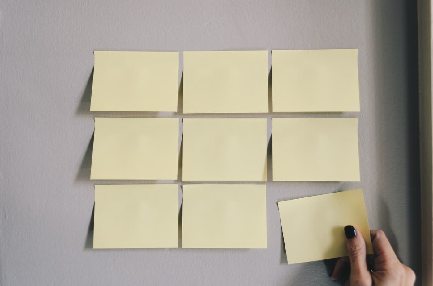 A grid of blank sticky notes on a wall with a hand placing the last one