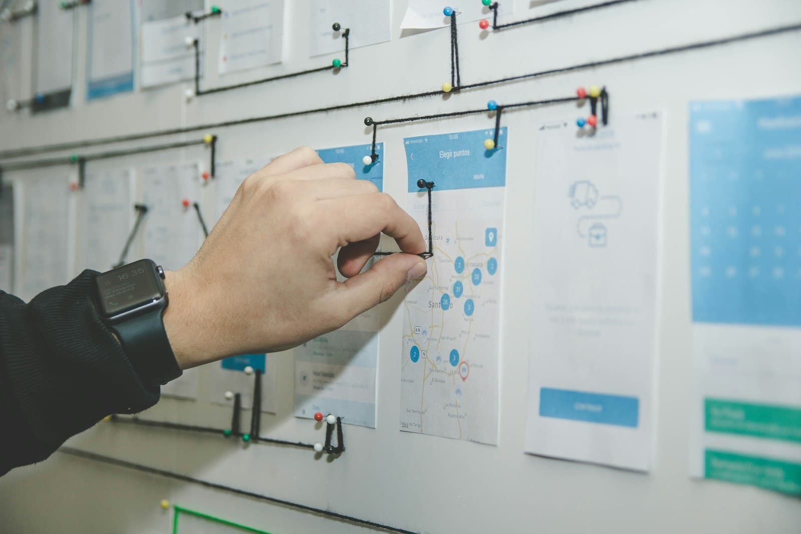 A hand connecting printed page layouts pinned across a wall with tape and markers