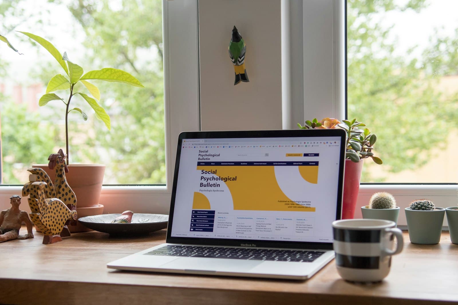 A laptop on a wooden desk by a window, showing a web page, with plants either side