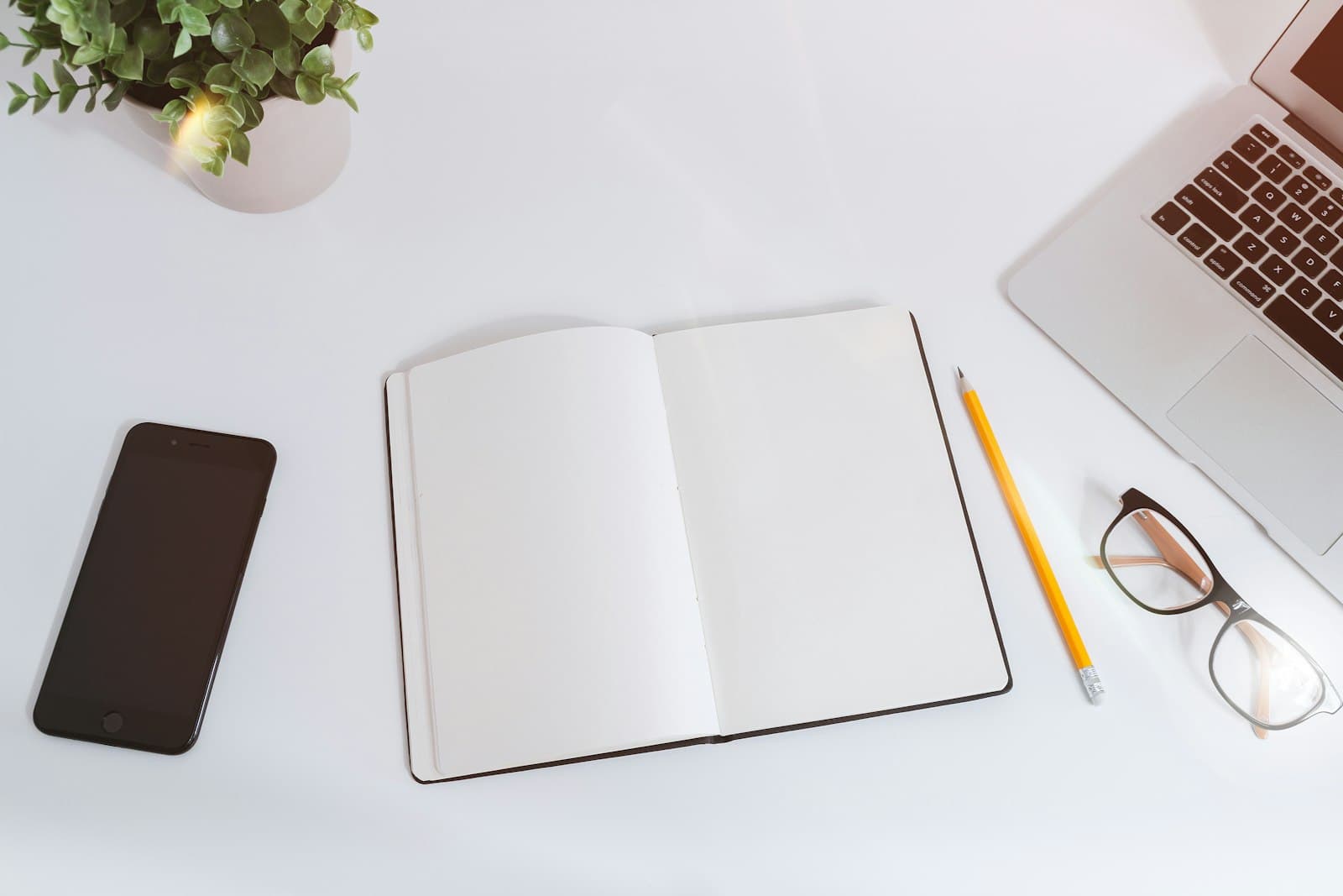An open blank notebook on a white desk beside a laptop, glasses and a small plant